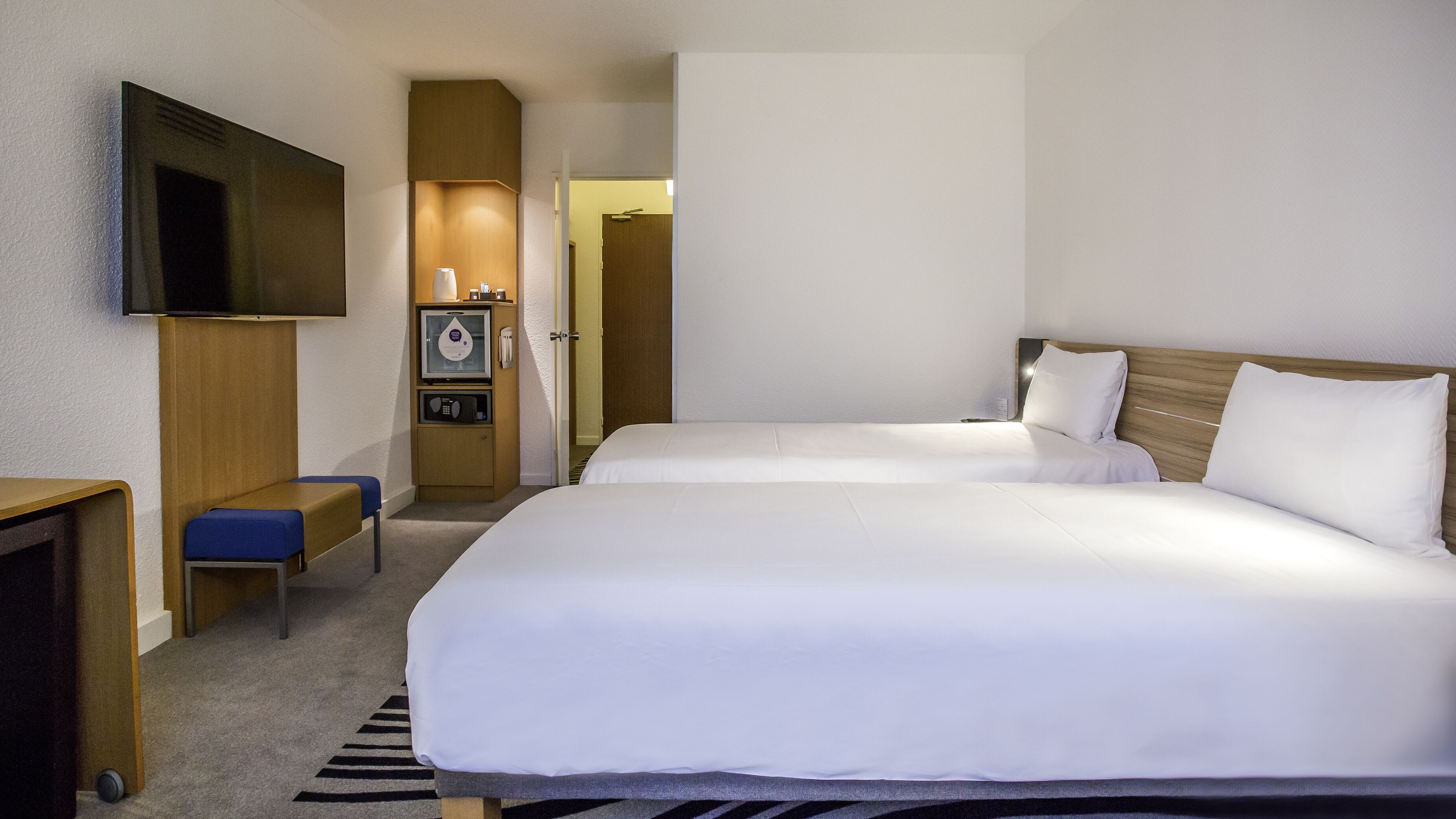 Habitación de hotel con dos camas, un televisor montado en la pared, un minibar y un escritorio con una silla.