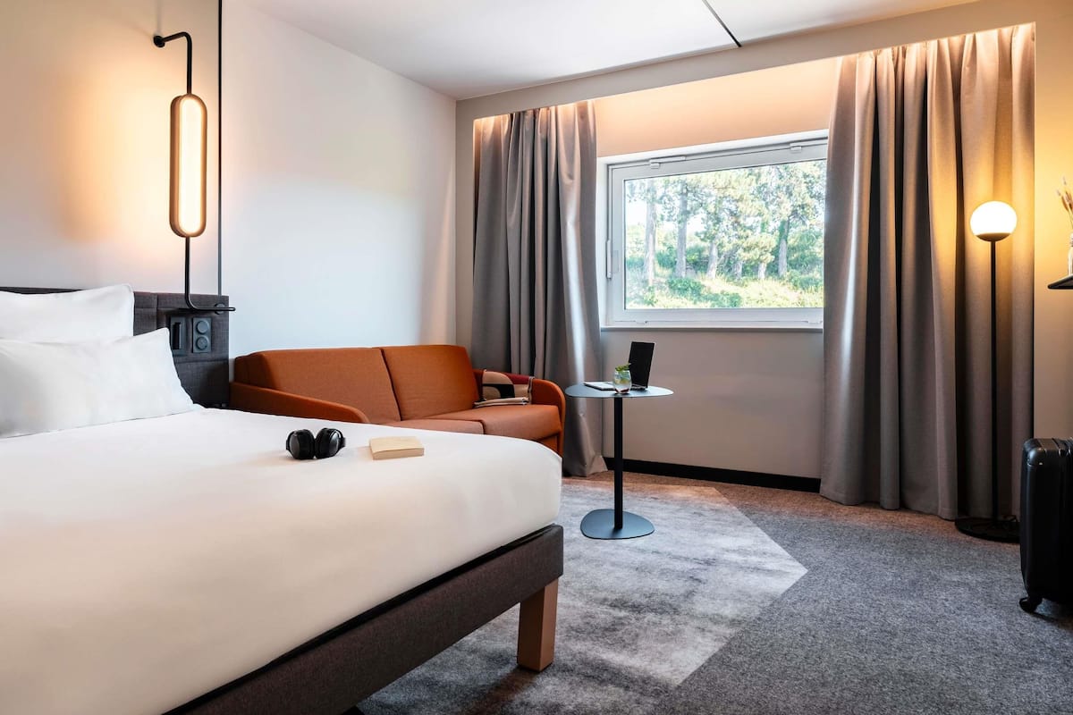 Ein modernes Hotelzimmer mit Bett, Sofa, einem kleinen Tisch, einer Lampe und einem Fenster mit Vorhängen.
