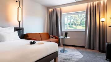A modern hotel room with a bed, a sofa, a small table, a lamp, and a window with curtains.