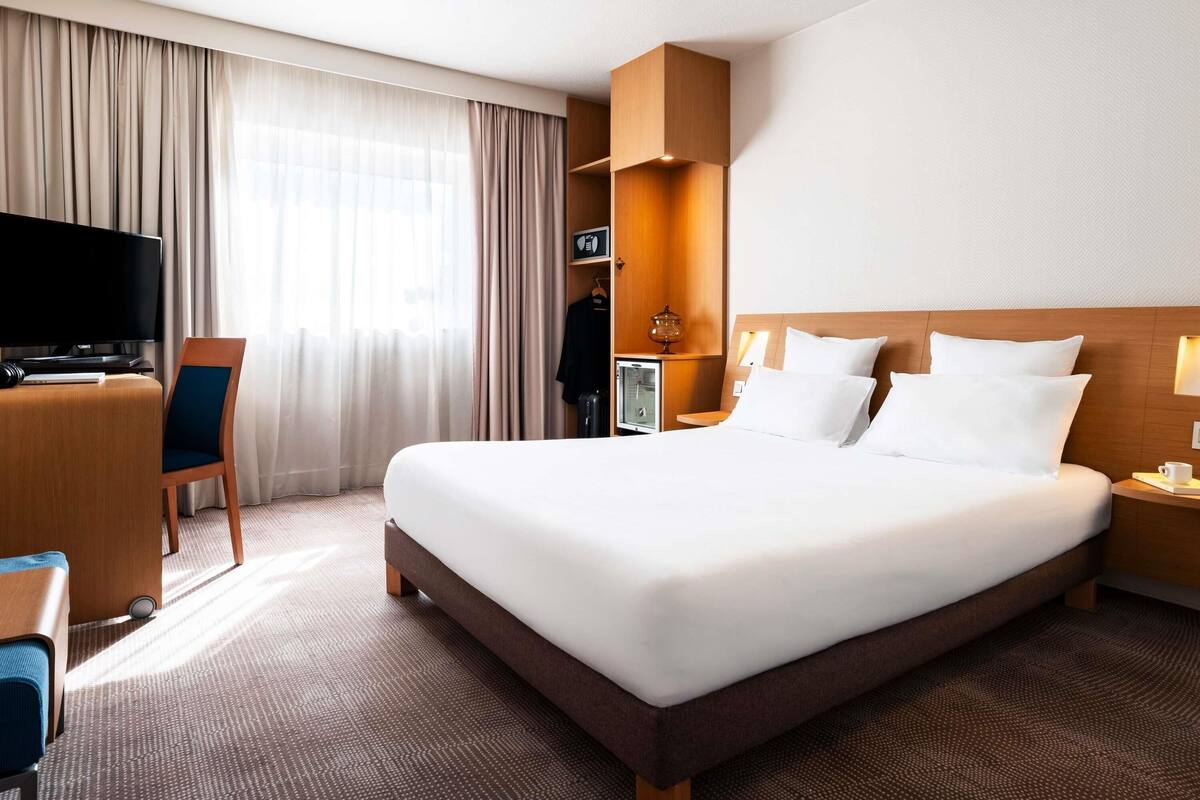 Ein Hotelzimmer mit einem großen Bett, einem Schreibtisch mit Fernseher, einem Sessel, einem Kleiderschrank und einem Nachttisch mit Lampe.