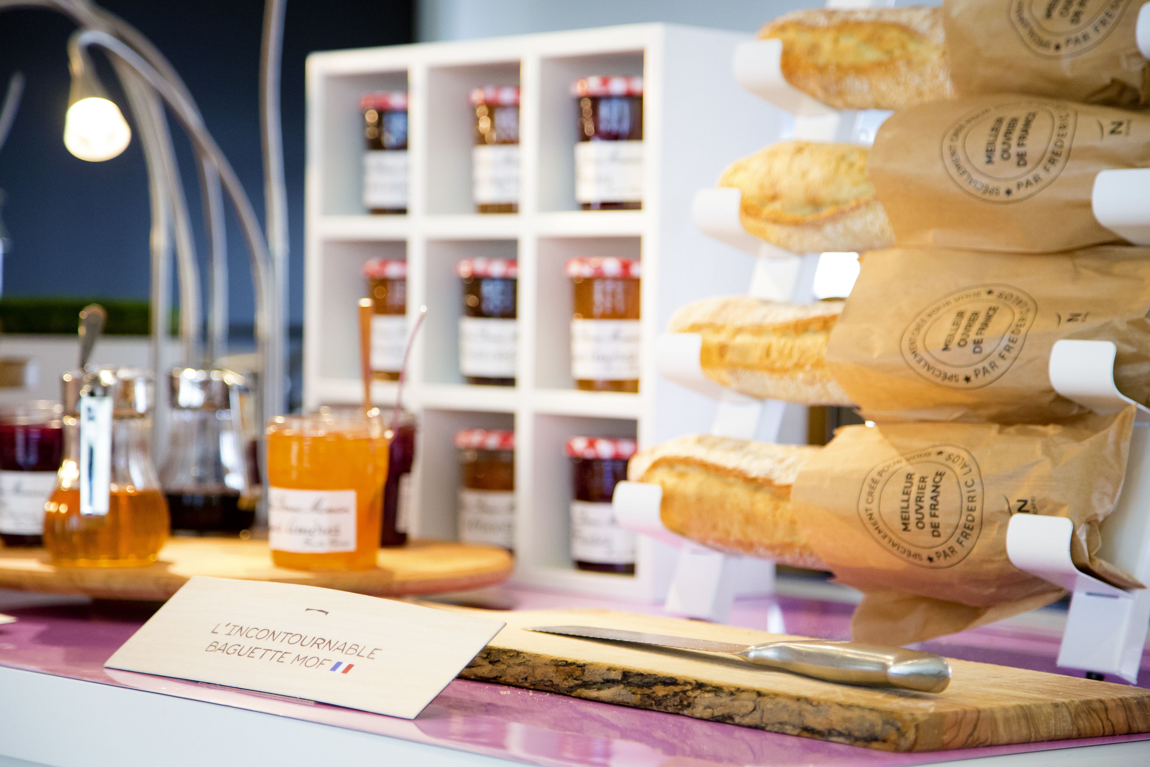 Une étalage de baguettes et de pots de miel est présenté sur un comptoir, avec une pancarte indiquant : « La baguette MOF, un incontournable ».