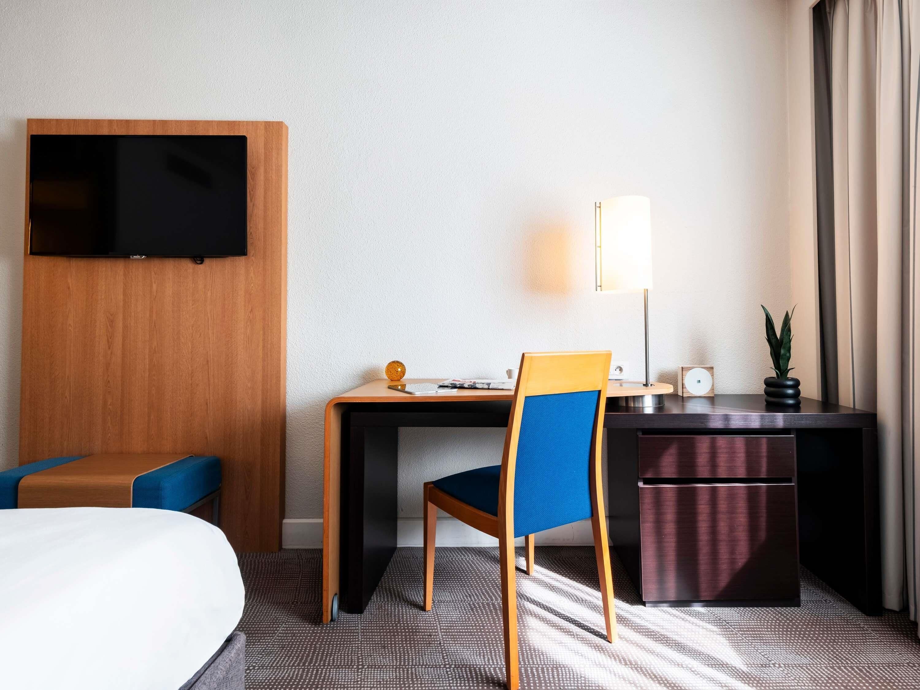Habitación de hotel con una cama, un escritorio con silla, un televisor montado en la pared, una lámpara, una planta pequeña y un minibar.