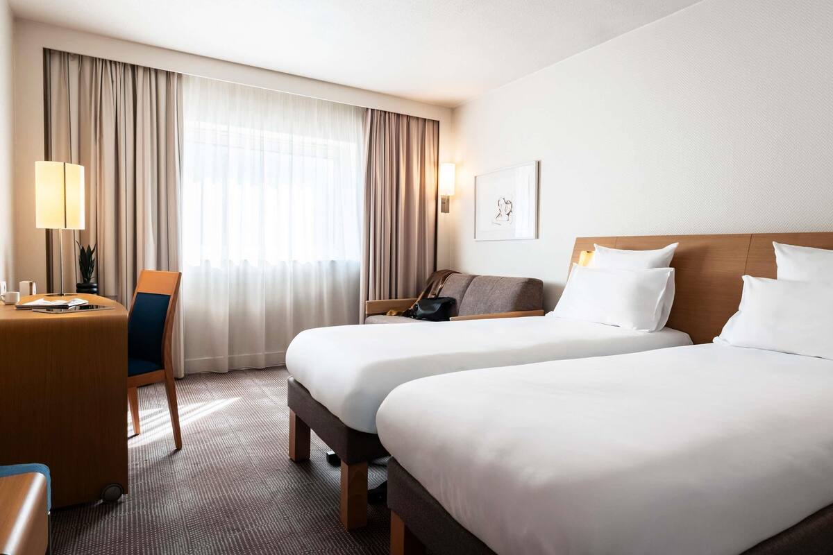 Ein Hotelzimmer mit zwei Betten, einem Schreibtisch, einem Stuhl, einer Lampe und einem gerahmten Bild an der Wand.
