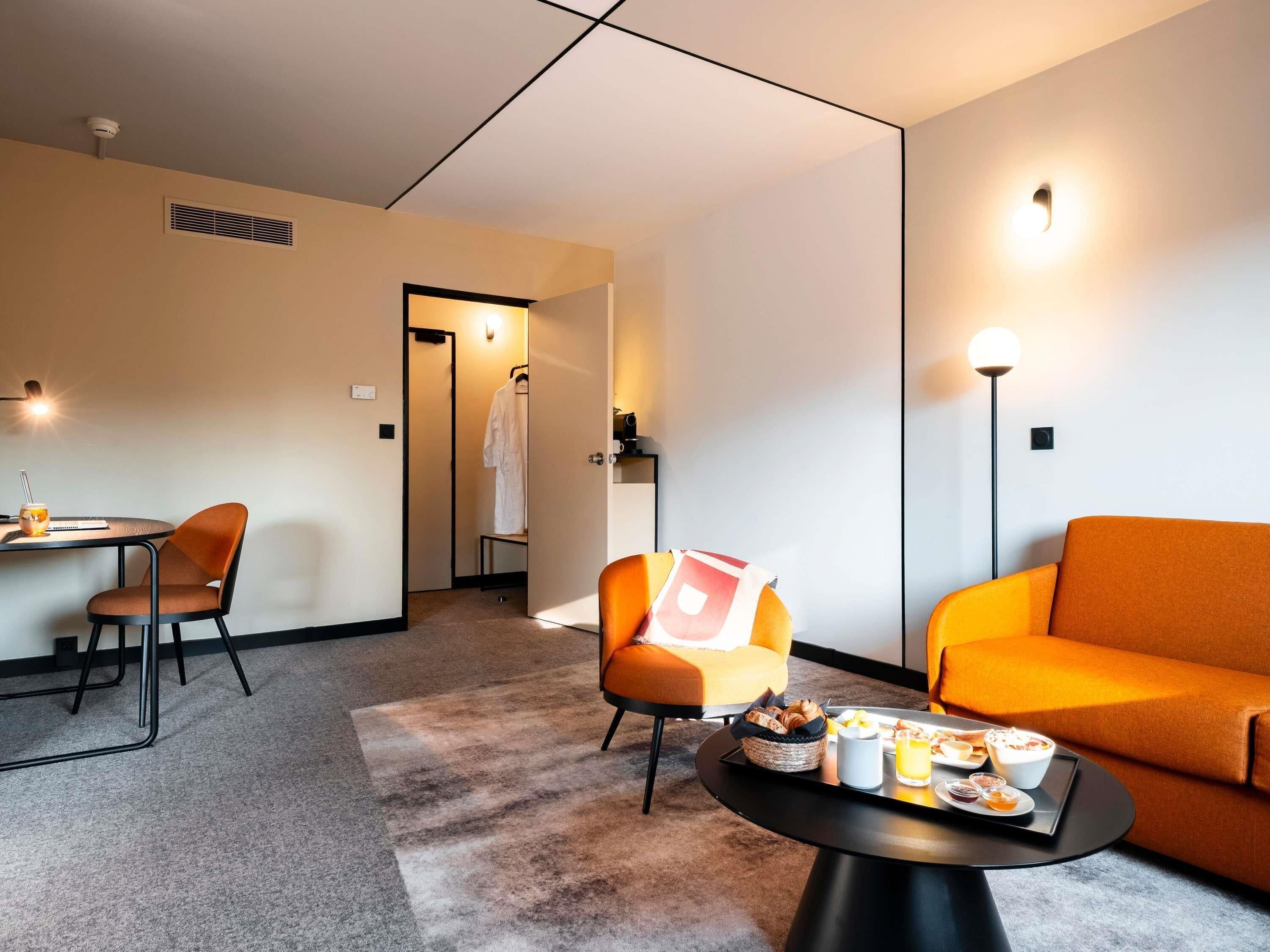 A modern hotel room with a dining area, a sitting area with a sofa, a table with breakfast, and a robe hanging in the doorway.