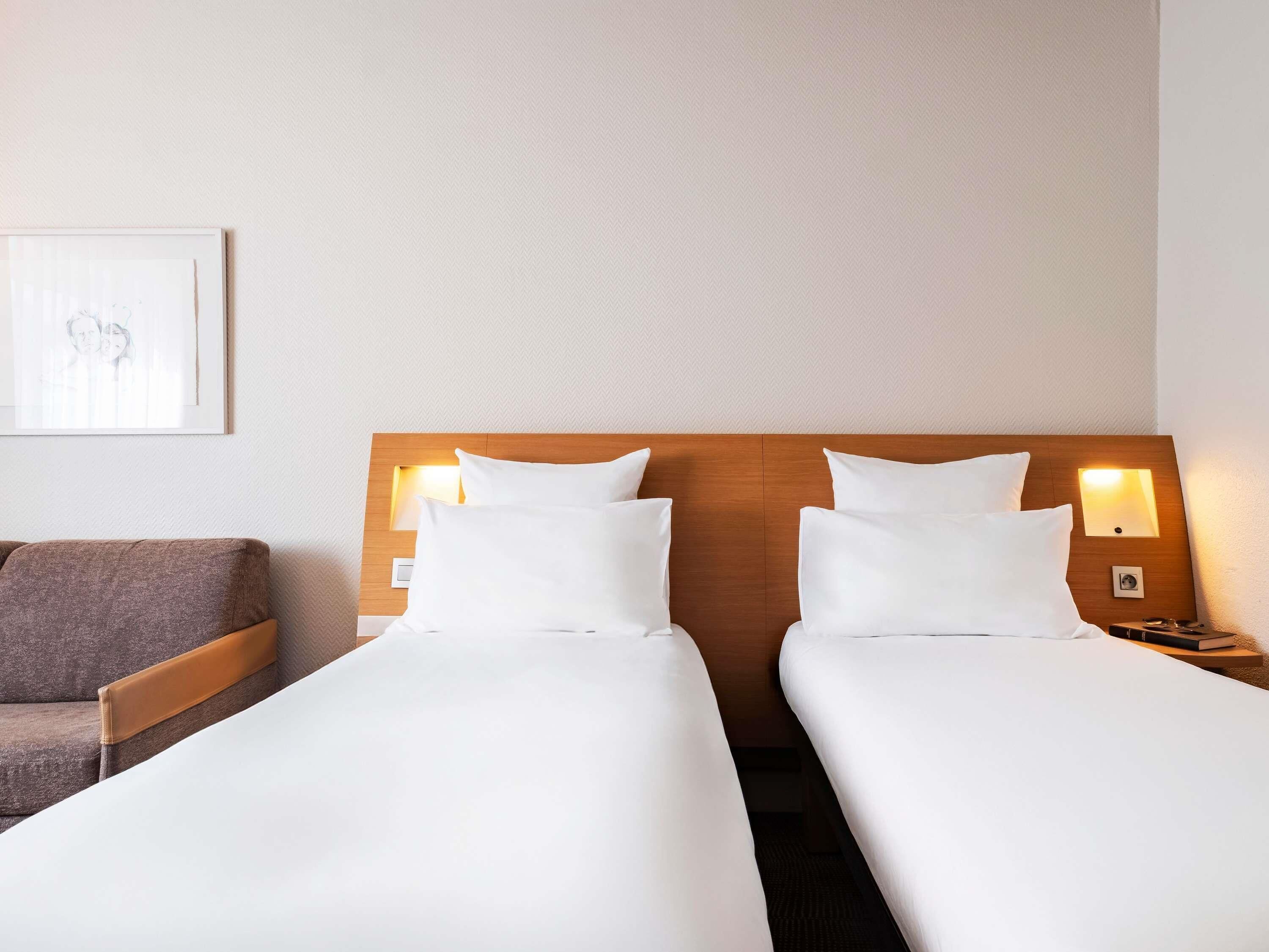 Una habitación de hotel con dos camas individuales, cabecero de madera, lámparas de noche, un cuadro enmarcado en la pared y un sofá gris.