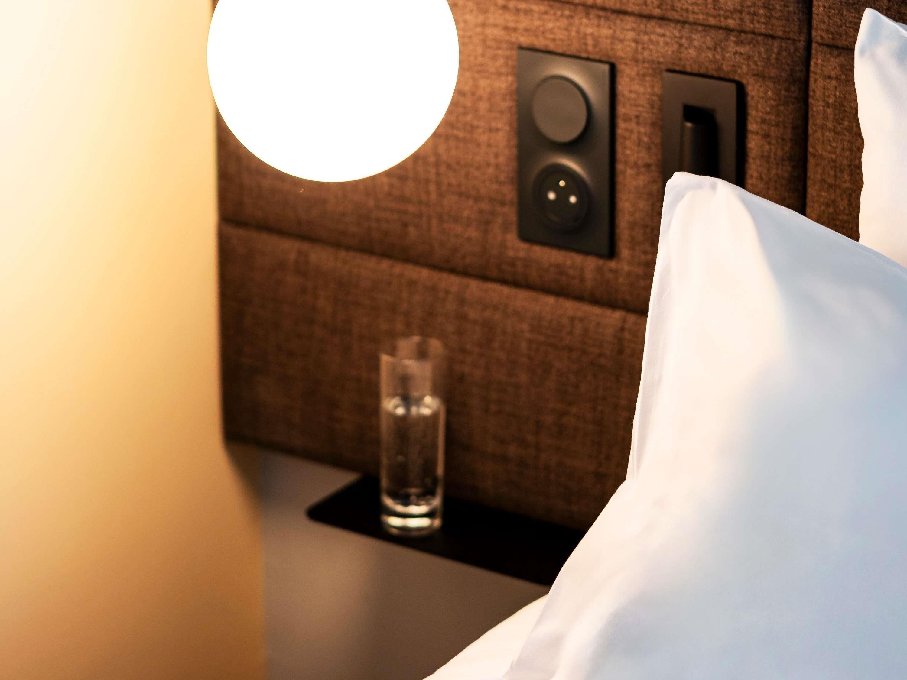 Habitación de hotel con una cama, mesita de noche, vaso de agua y panel de control en la pared.