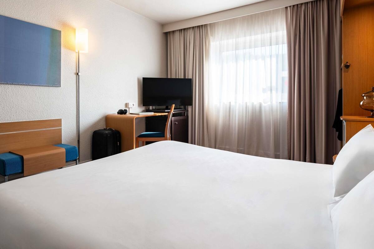 Ein Hotelzimmer mit einem großen Bett, einem Schreibtisch mit Fernseher, einem Sessel, einem Koffer und einer Lampe.