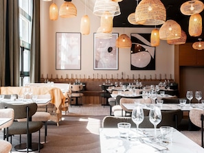 A restaurant interior with multiple dining tables, wicker pendant lights, and framed artwork on the walls.