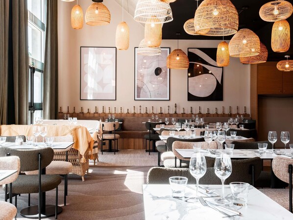 A restaurant interior with multiple dining tables, wicker pendant lights, and framed artwork on the walls.