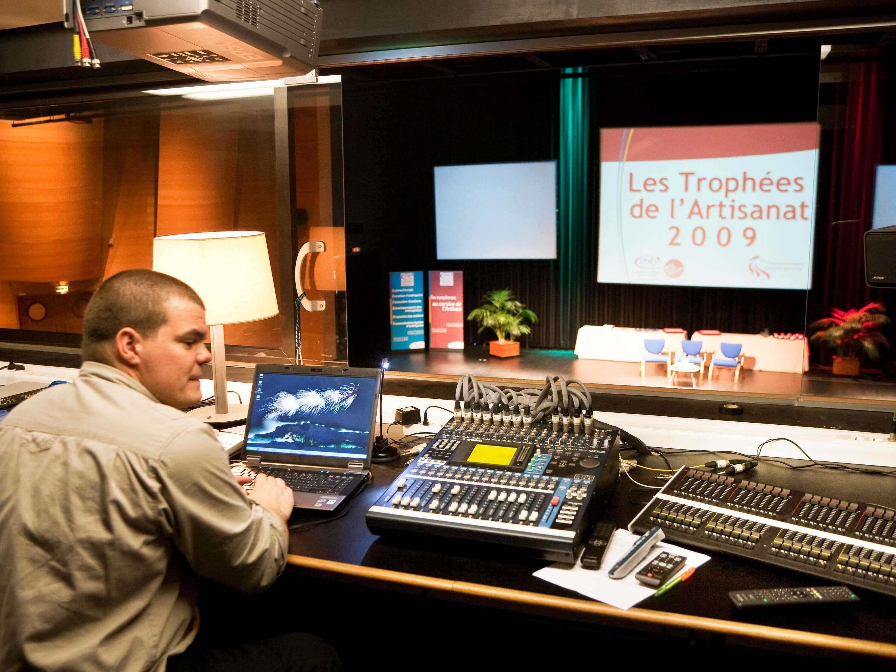 Un homme travaille à un bureau équipé de matériel audio, d’un ordinateur portable et d’un écran de présentation affichant le texte « Les Trophées de l’Artisanat 2009 ».