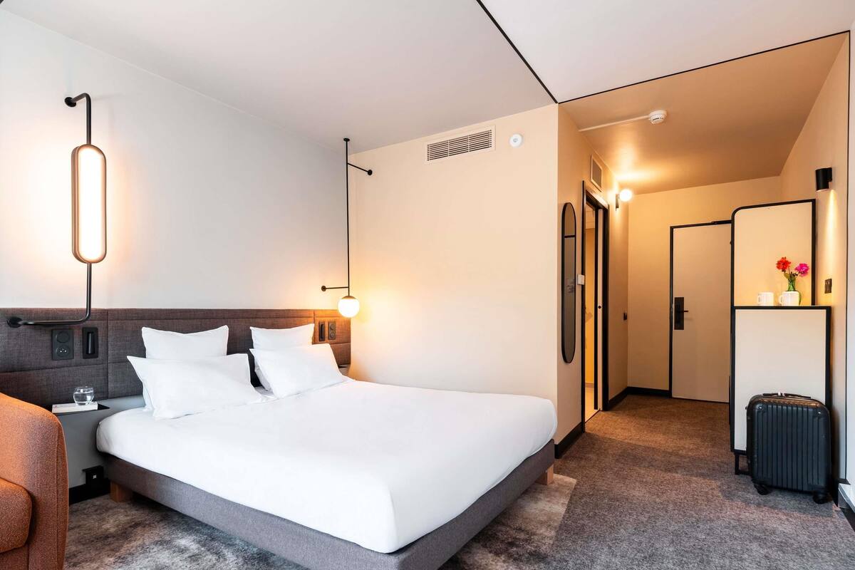 Ein modernes Hotelzimmer mit einem großen Bett, Nachttischen, einem Kleiderschrank und einem durchgehenden Türrahmen einsehbarer Badezimmer.