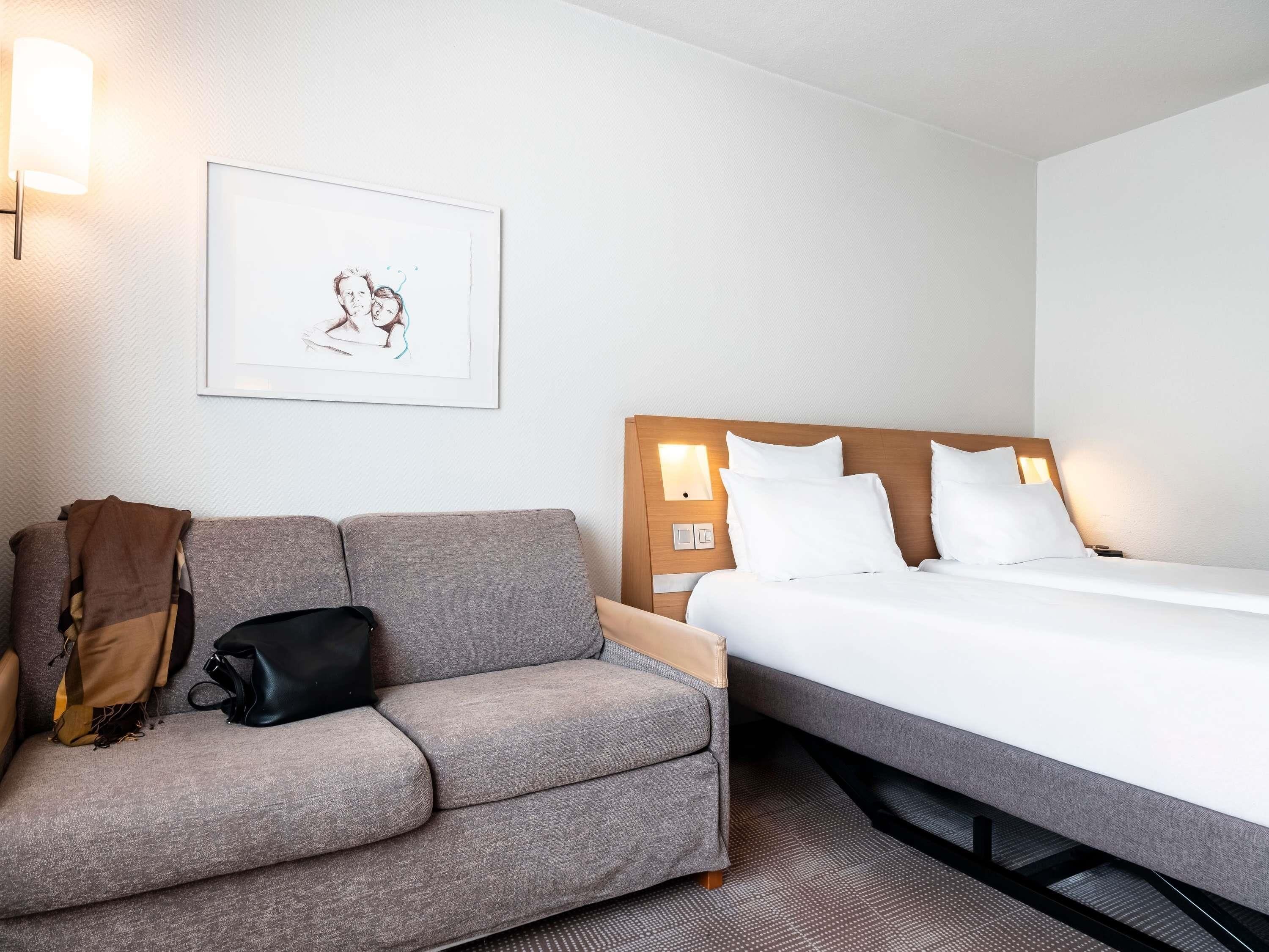 Habitación de hotel con un sofá gris, una cama con ropa blanca, un cuadro en la pared y una mesita de noche con una lámpara.