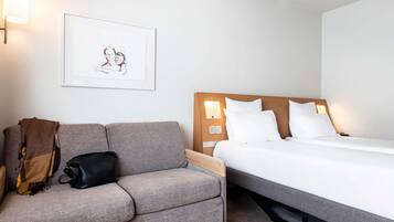 Habitación de hotel con un sofá gris, una cama con ropa blanca, un cuadro en la pared y una mesita de noche con una lámpara.