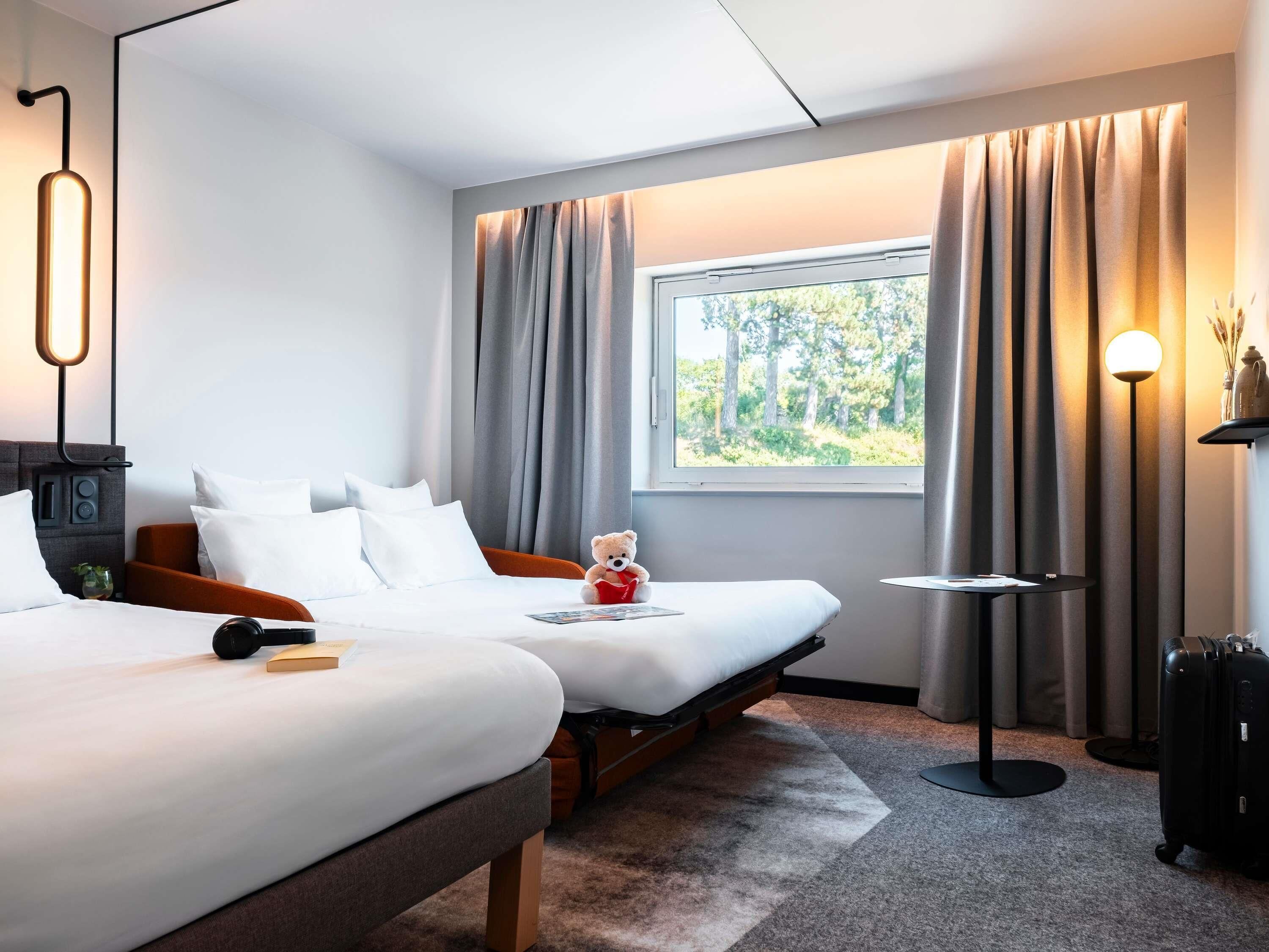 A hotel room with two beds, a window with curtains, a bedside table, a lamp, and a teddy bear on the left bed.