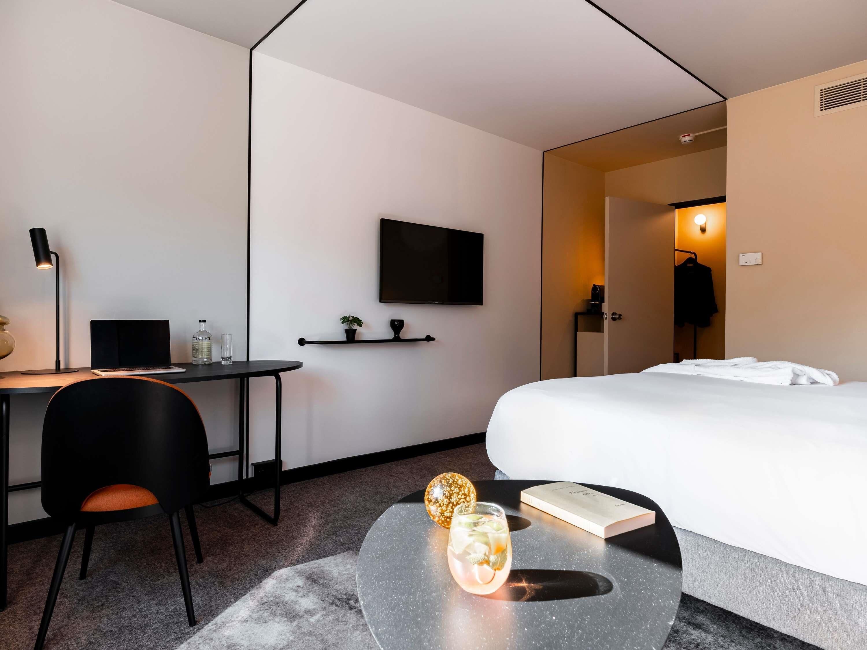 Una habitación de hotel moderna con cama, escritorio, silla y una pequeña mesa con una bebida y un libro.
