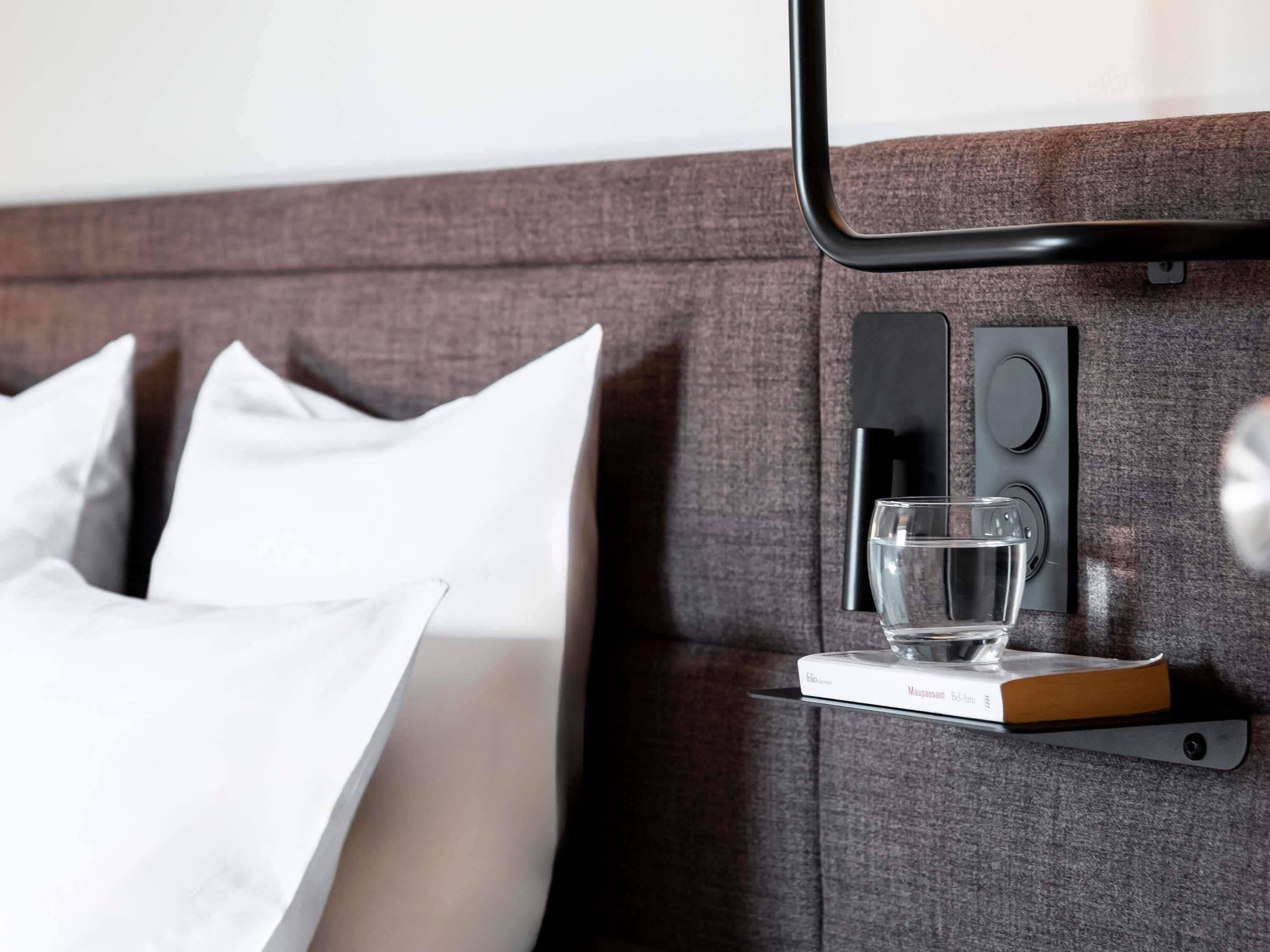 Una cama bien tendida con almohadas blancas, una mesita de noche con un vaso de agua, un libro y un altavoz.