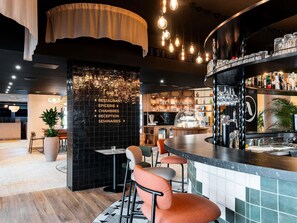 A modern bar area with a black tiled wall, a wooden reception desk, and a bar counter with various bottles and glasses.