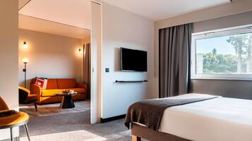 Une chambre d’hôtel avec un lit, un téléviseur fixé au mur, un canapé, une table basse et une vue sur les arbres par la fenêtre.