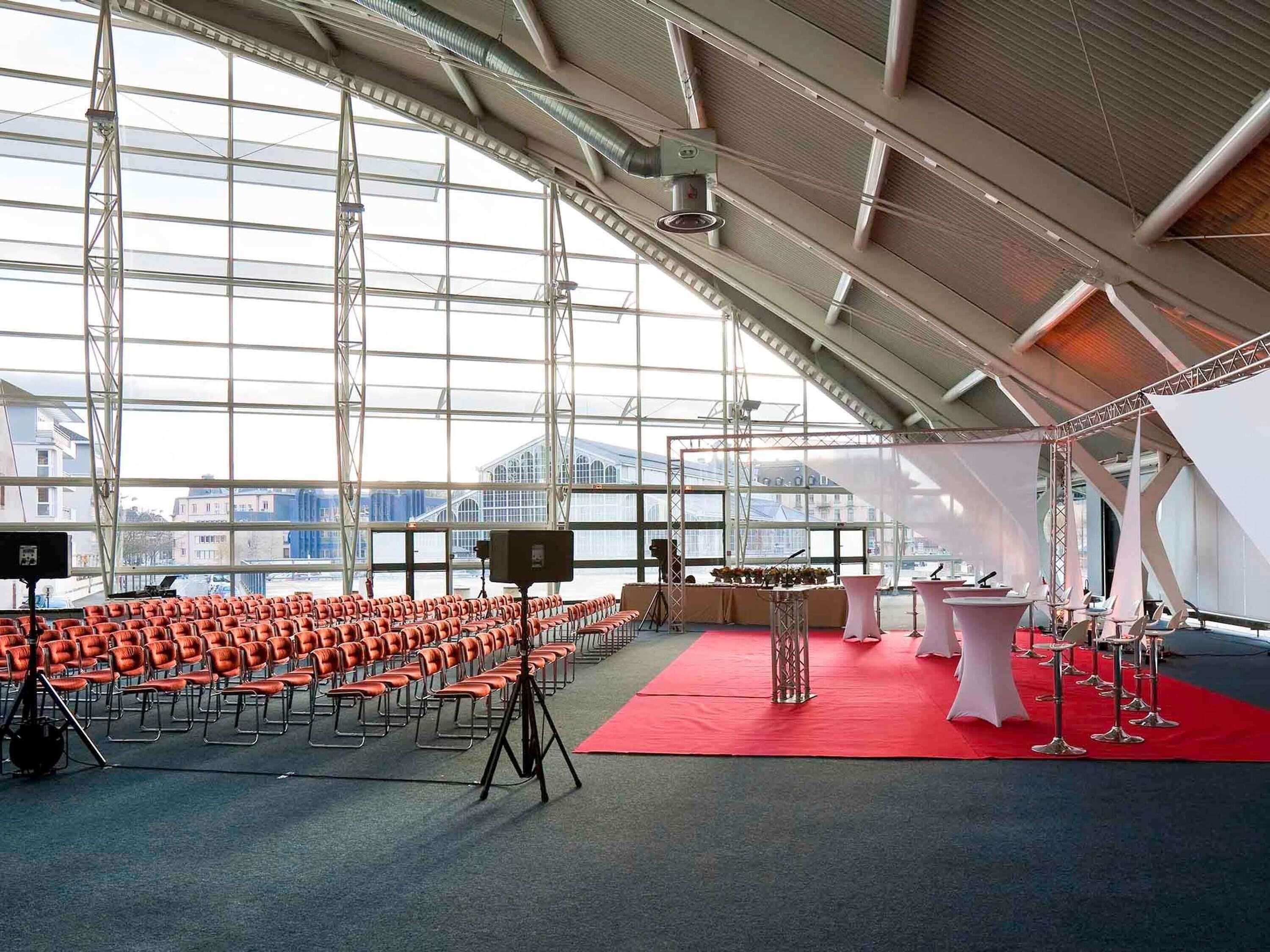 A large indoor venue with rows of chairs, a stage with a red carpet, and a backdrop.