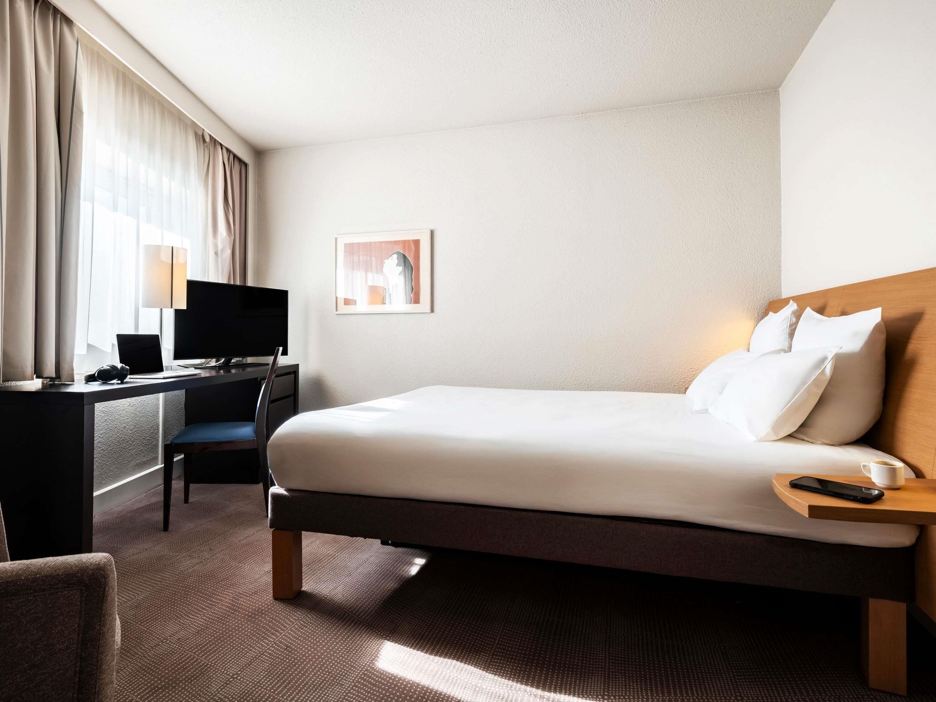 A hotel room with a bed, a desk with a computer, a chair, a TV, and a picture on the wall.