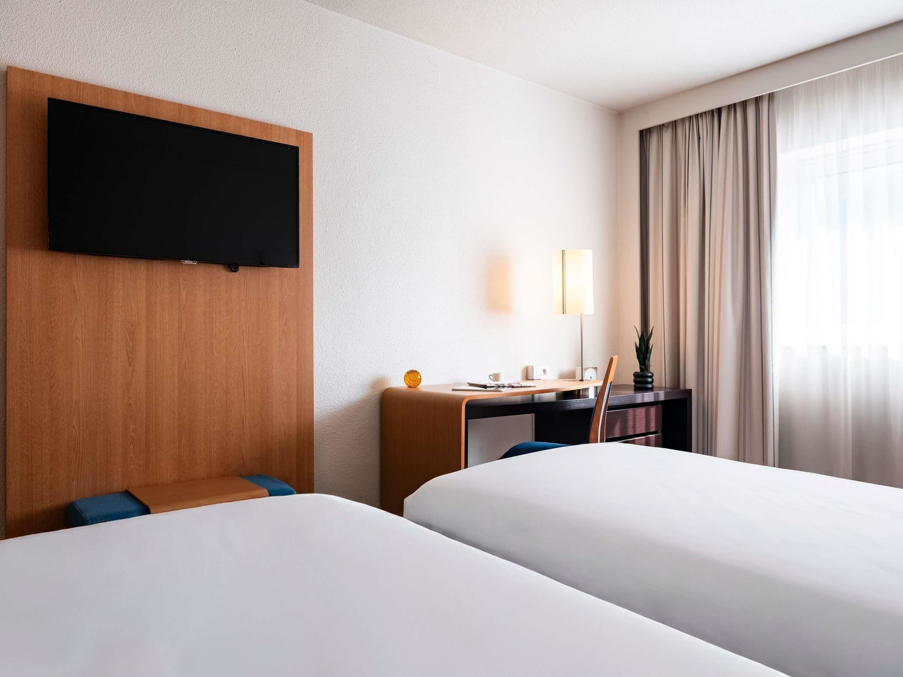 A hotel room with two beds, a desk with a lamp, a TV mounted on the wall, and a window with curtains.