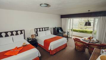 A hotel room with two beds, a desk with a computer, a dining table with breakfast, and a view of greenery.