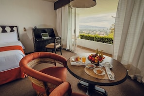 A hotel room with a bed, a desk, a chair, a balcony with a view, and a breakfast table with fruit and drinks. - Hotel Victoria Oaxaca (Oaxaca)
