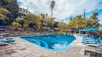 A swimming pool with lounge chairs and umbrellas, surrounded by palm trees and a building in the background.