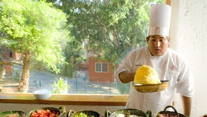 A chef holding a large yellow dish, with various bowls of food in front of a window overlooking a street. - Hotel Victoria Oaxaca (Oaxaca)