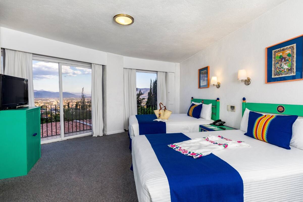 Habitación de hotel con dos camas, televisión, un armario verde y balcón con vistas a la ciudad.