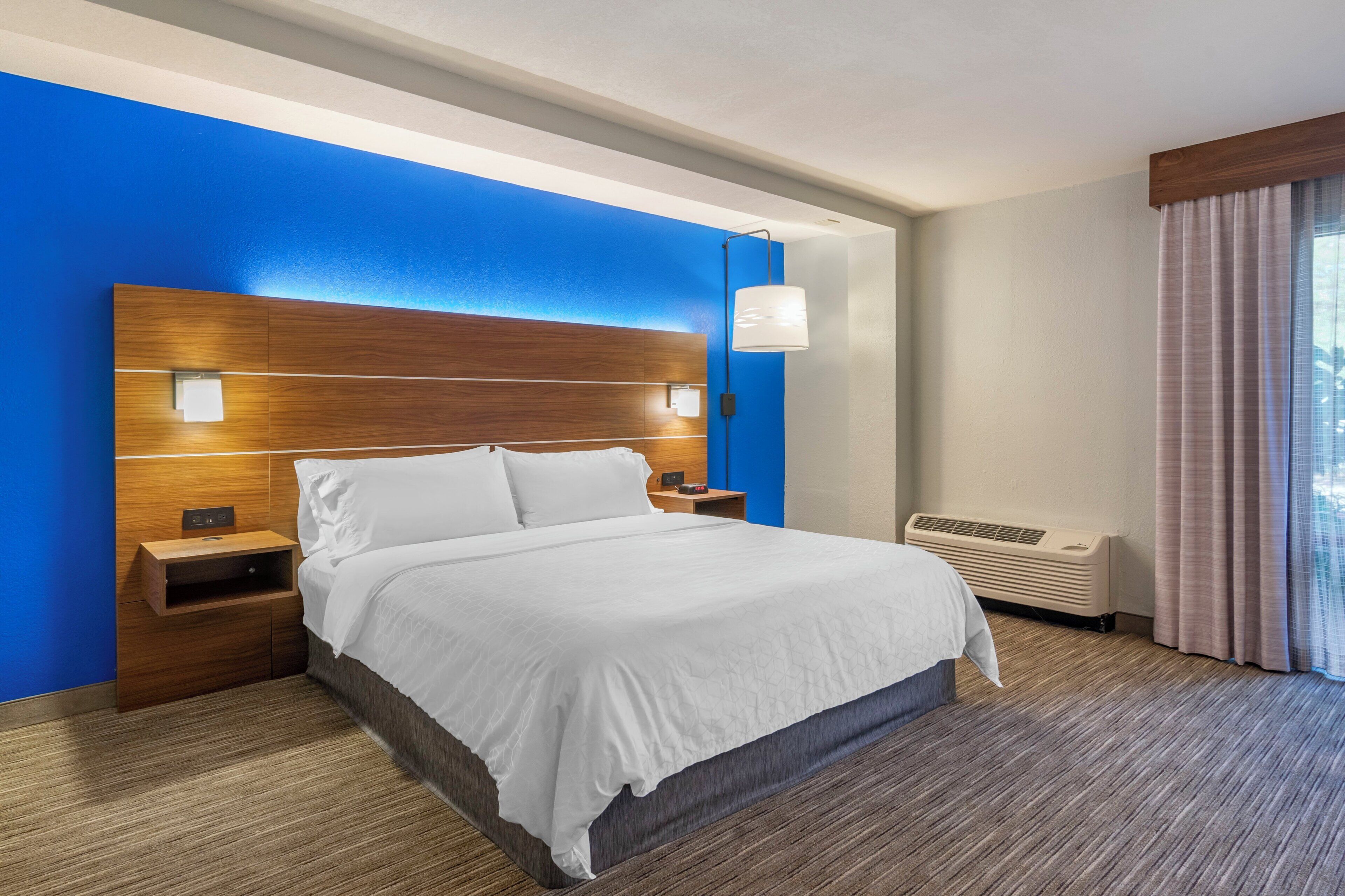 A hotel room with a large bed, a nightstand, a wall-mounted lamp, and an air conditioning unit.