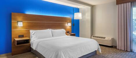 A hotel room with a large bed, a nightstand, a wall-mounted lamp, and an air conditioning unit.