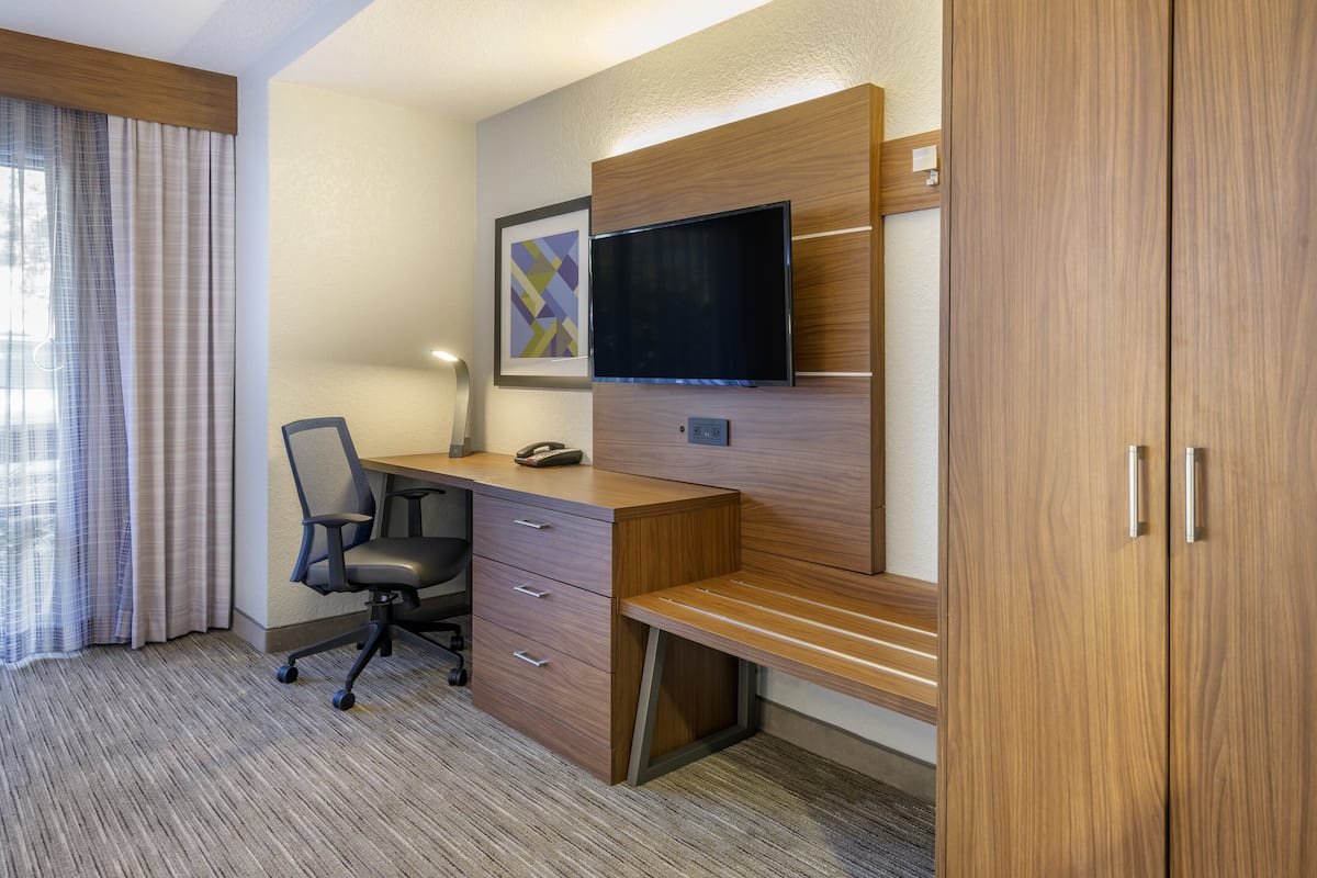 A hotel room with a wooden desk, a chair, a flat-screen TV mounted on the wall, and a large wardrobe.