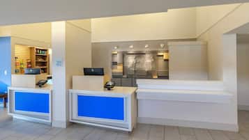 Zona de recepción de un hotel con mostrador de registro, quiosco de auto-servicio y una pequeña área de cocina.