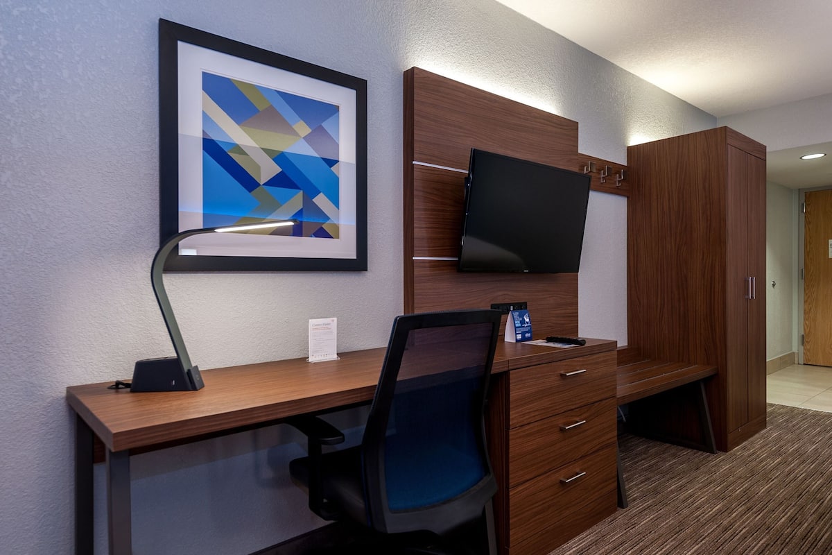A hotel room with a wooden desk, a black office chair, a framed abstract artwork, a flat-screen TV, and a wardrobe.