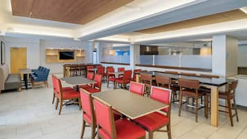 A modern restaurant with multiple dining tables and chairs, a kitchen area, and a flat-screen TV mounted on the wall.