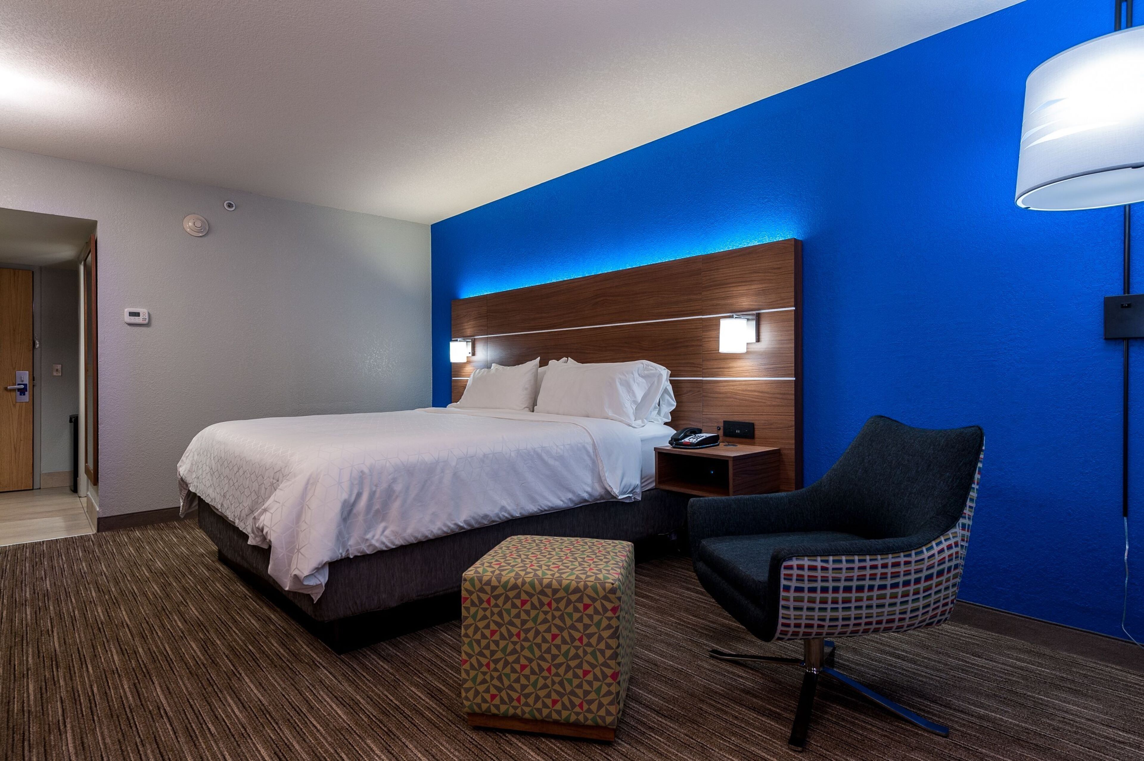 A hotel room with a bed, a chair, a nightstand, and a wall-mounted lamp.