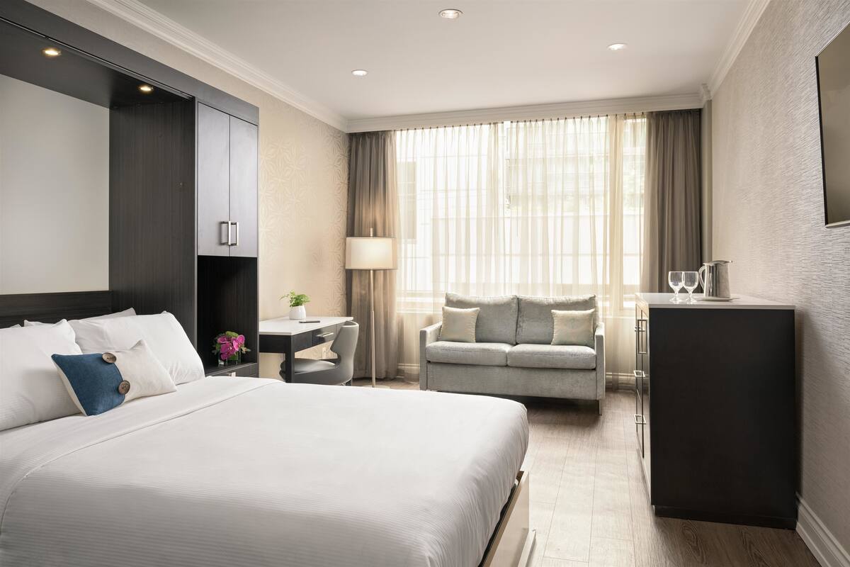A hotel room with a large bed, a desk, a sofa, and a minibar.