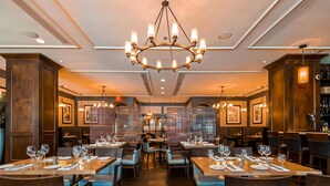 A restaurant interior with multiple dining tables set with glasses and cutlery, a large chandelier, and framed pictures on the walls.
