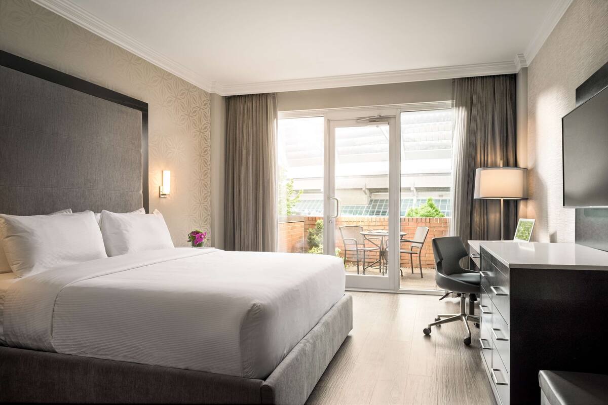 A hotel room with a large bed, a desk with a chair, a TV, and a balcony with a table and chairs.