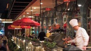 A restaurant with outdoor seating, red umbrellas, and a chef preparing food.