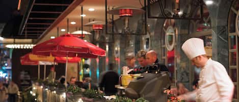 Un ristorante con posti a sedere all'aperto, ombrelloni rossi e uno chef che prepara dei piatti.