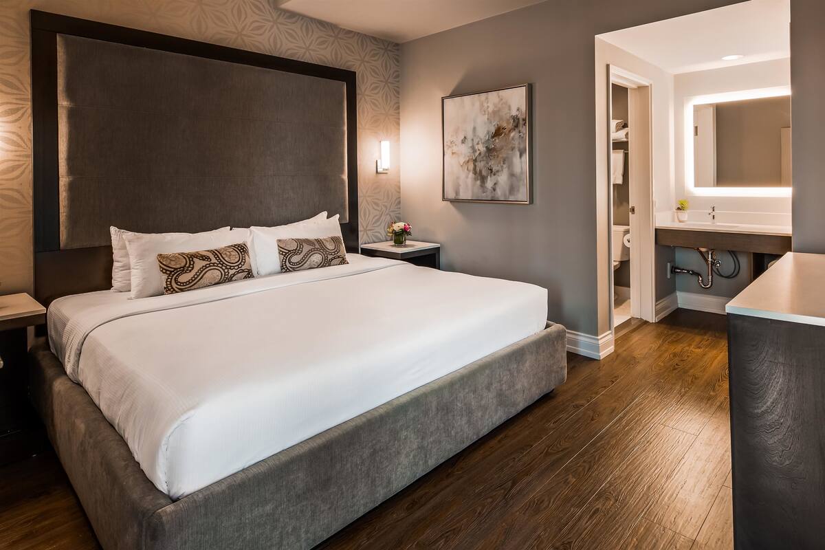 A modern hotel room with a large bed, a bedside table, a nightstand, a bathroom with a sink and mirror, and a wooden floor.