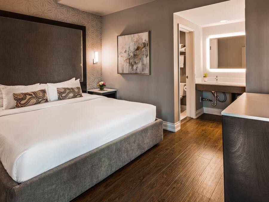 Una habitación de hotel moderna con una cama grande, una mesita de noche, un tocador, un baño con lavabo y espejo, y suelo de madera.
