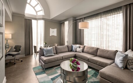 A modern living room with a sectional sofa, a glass coffee table with flowers, a desk with a chair, and large windows with curtains.