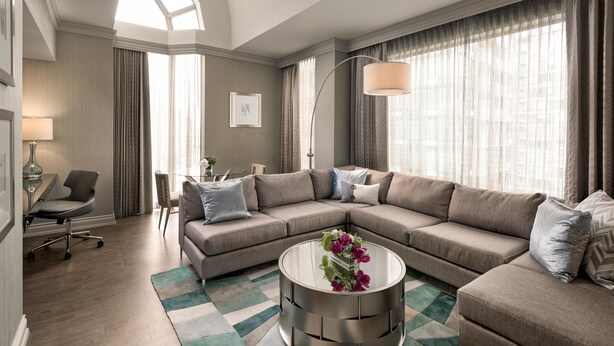 A modern living room with a sectional sofa, a glass coffee table with flowers, a desk with a chair, and large windows with curtains.