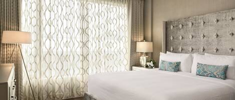 A hotel room with a large bed, a nightstand with a lamp, and a window with patterned curtains.
