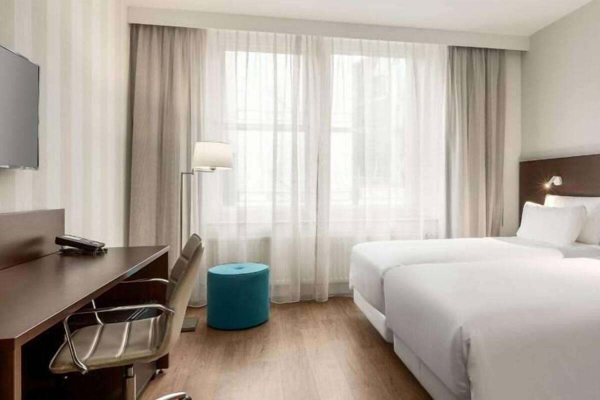 A hotel room with a bed, a desk, a chair, a lamp, and a window with curtains.