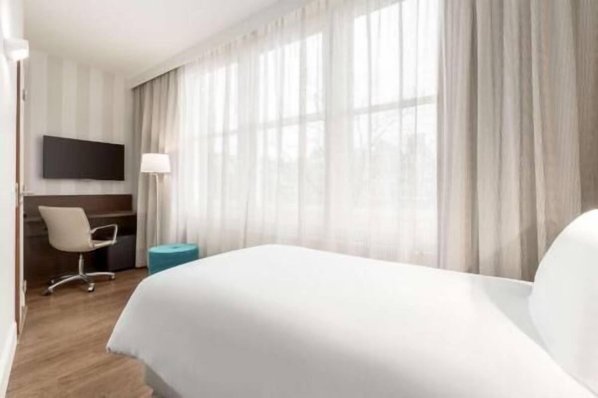 A hotel room with a large bed, a desk with a chair, a flat-screen TV, and sheer curtains.