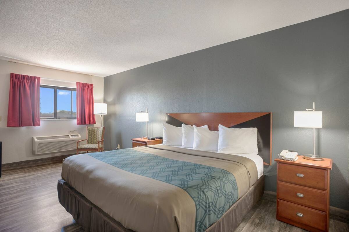 Una habitación de hotel con una cama, dos mesitas de noche, una silla, una ventana con cortinas y un radiador.